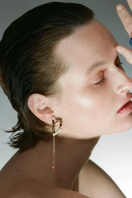 Close-up beauty shot of a woman with short hair and a gold drop earring with gemstones, gazing downward in soft studio lighting.