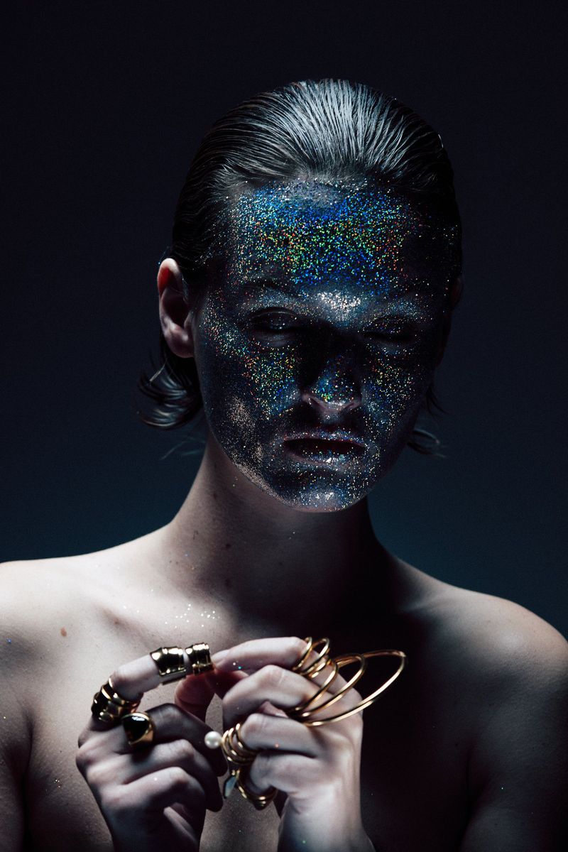 Close-up portrait of a person with glitter on their face and gold rings. Moody lighting illuminates their face and jewelry.