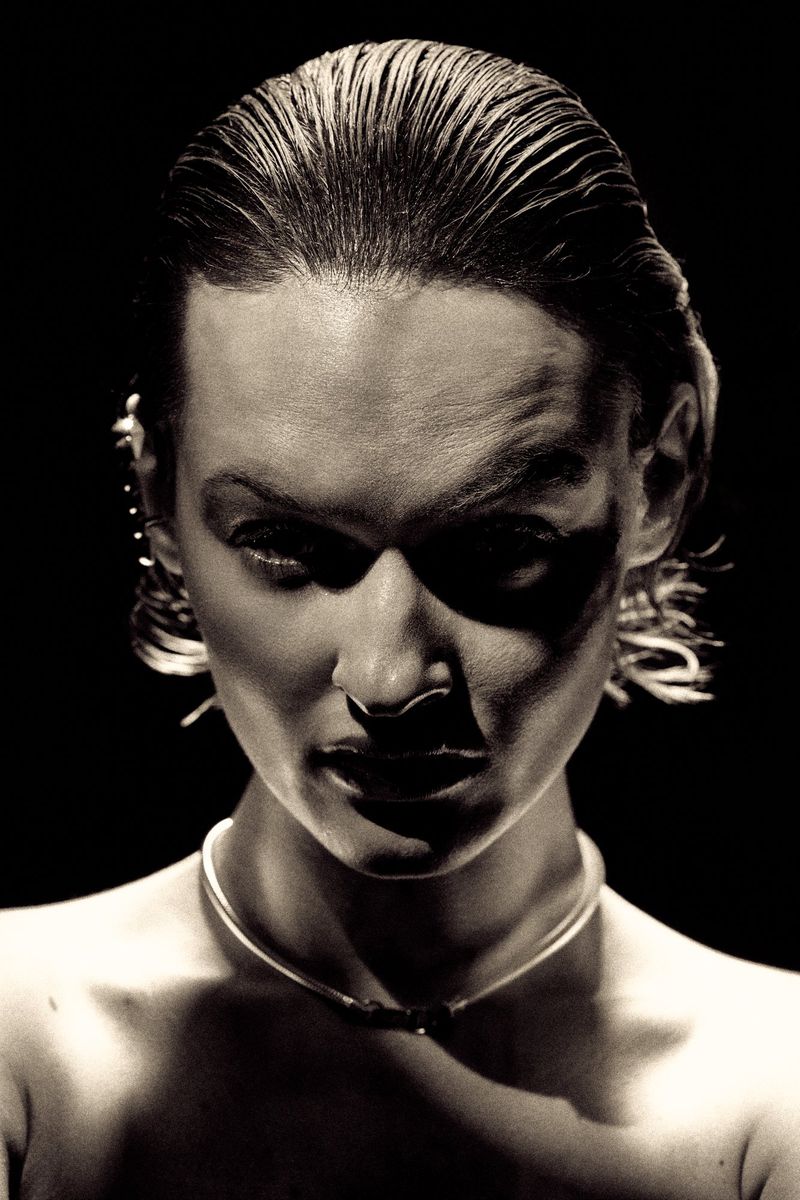 Close-up portrait of a fair-skinned adult female with slicked-back hair, wearing a thin silver necklace, set against a stark black backdrop.