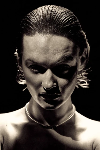 Close-up portrait of a fair-skinned adult female with slicked-back hair, wearing a thin silver necklace, set against a stark black backdrop.