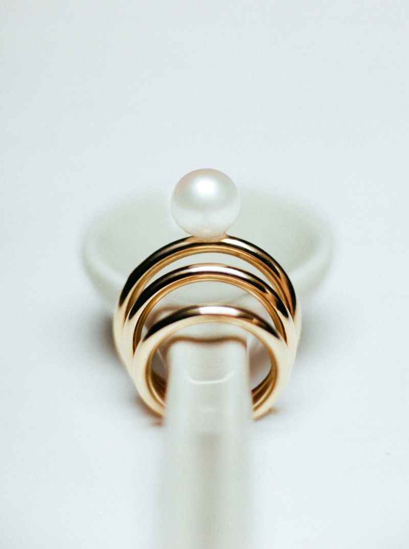 Close-up of a gold ring with a pearl on top, resting on a white ceramic spoon against a stark white background. Jewellery fashion.