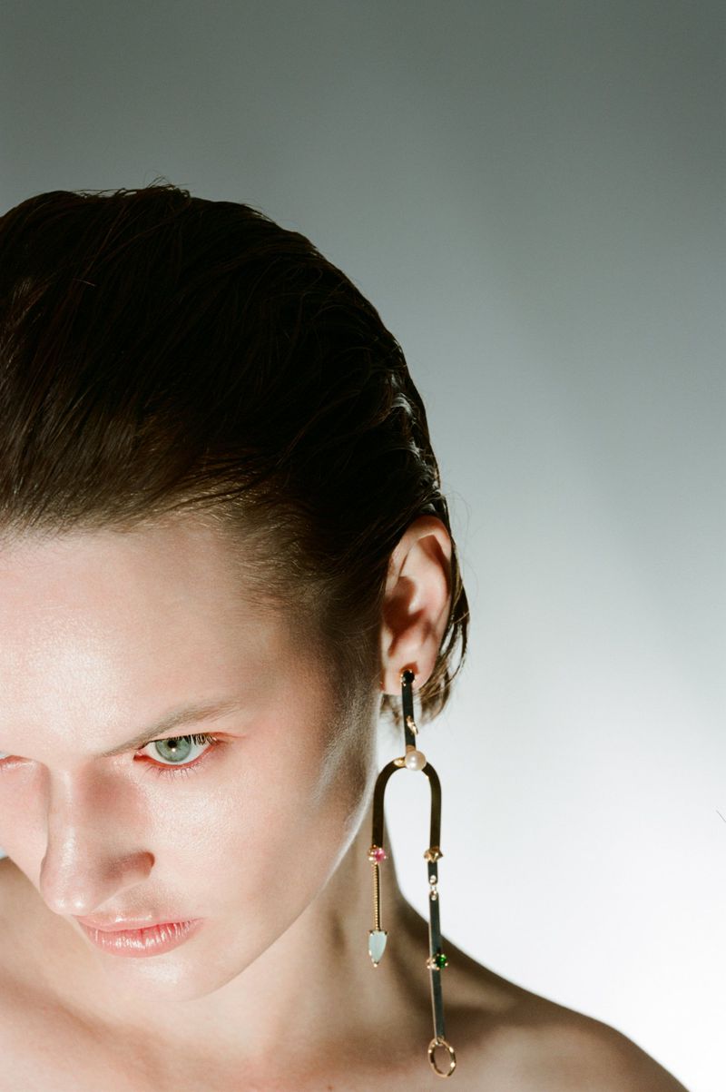 Close-up studio shot of a woman with slicked-back hair wearing a long, dangly gold earring with colorful gemstone accents.