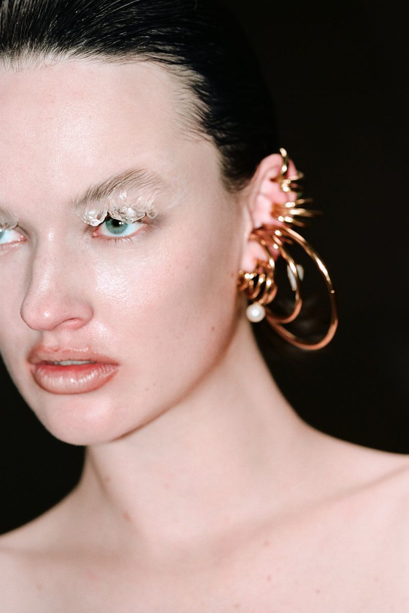 Close-up beauty shot of a woman with pale skin wearing statement gold jewelry and dramatic white eyeshadow against a dark background.