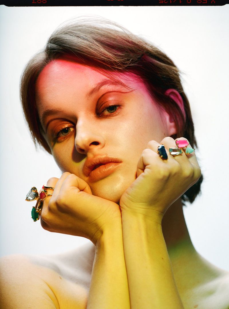 Close-up portrait of a woman with short brown hair, her face resting on her hands adorned with gemstone rings, against a white backdrop.