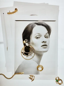 Black and white portrait photograph of a woman overlaid with layered gold hoop earrings and jewelry on sheets of white paper.