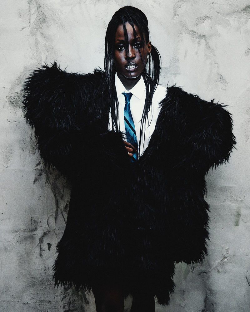 A person with braided hair wears a black faux fur coat over a white shirt and patterned tie against a grey textured wall backdrop.