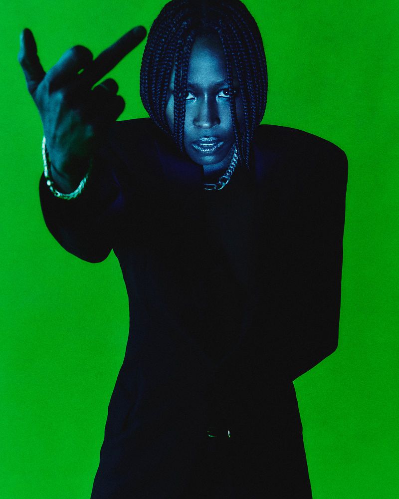Portrait of an adult with braided hair and a black blazer making an obscene gesture against a vivid green background in a studio shot.