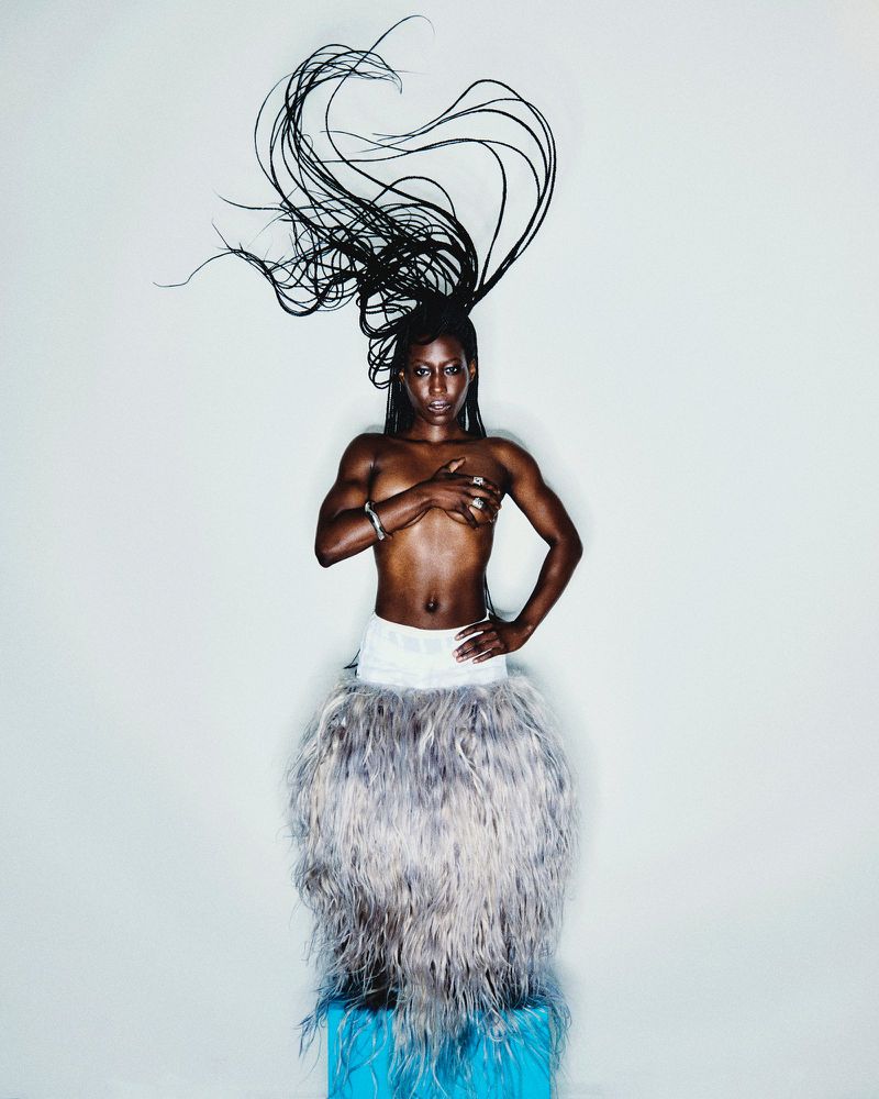 Striking portrait of Black woman with elaborate braided hair and grey feathered skirt against a plain white backdrop in a studio setting.