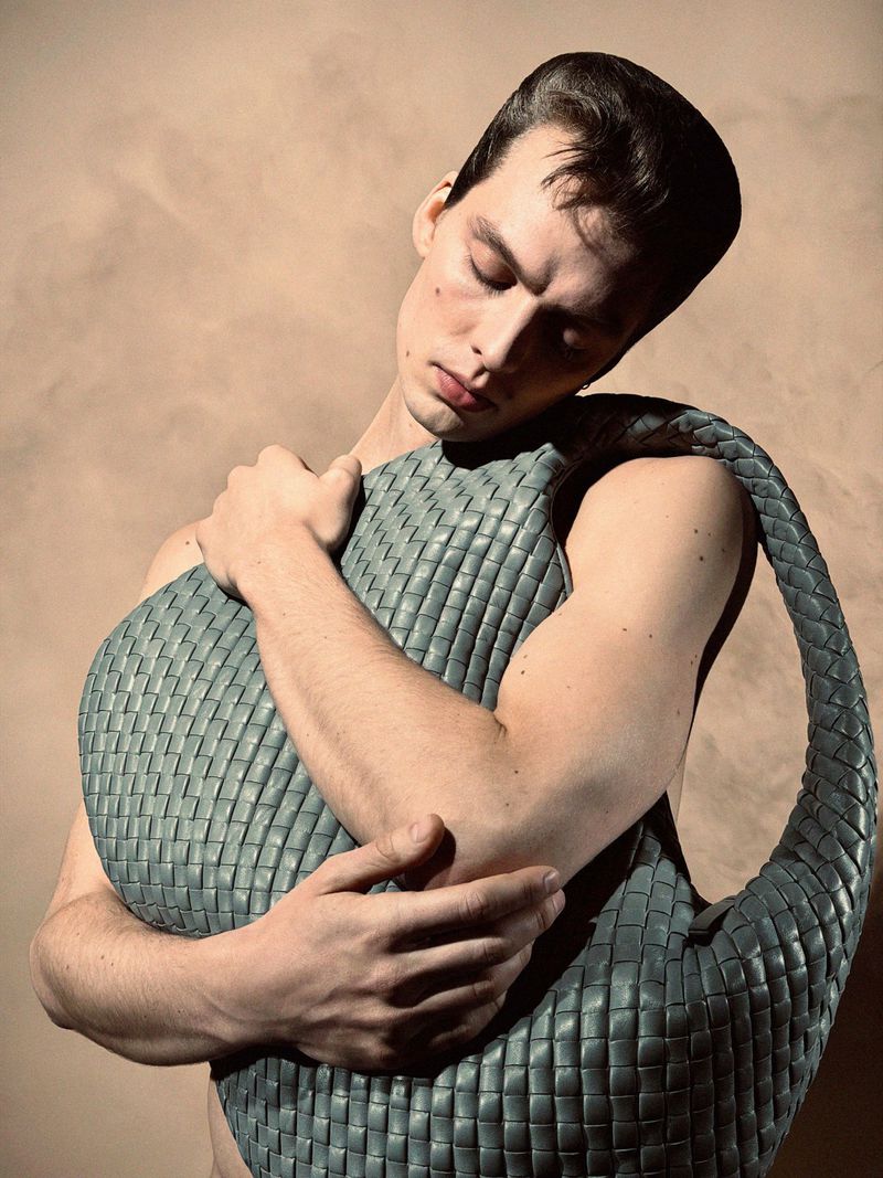 Close-up of a shirtless man with dark hair holding a woven blue leather bag against a tan background in a studio shot.