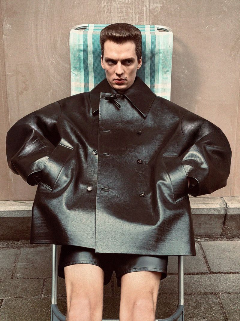Adult male in black leather coat and shorts sits on a light blue striped beach chair against a textured beige outdoor background.