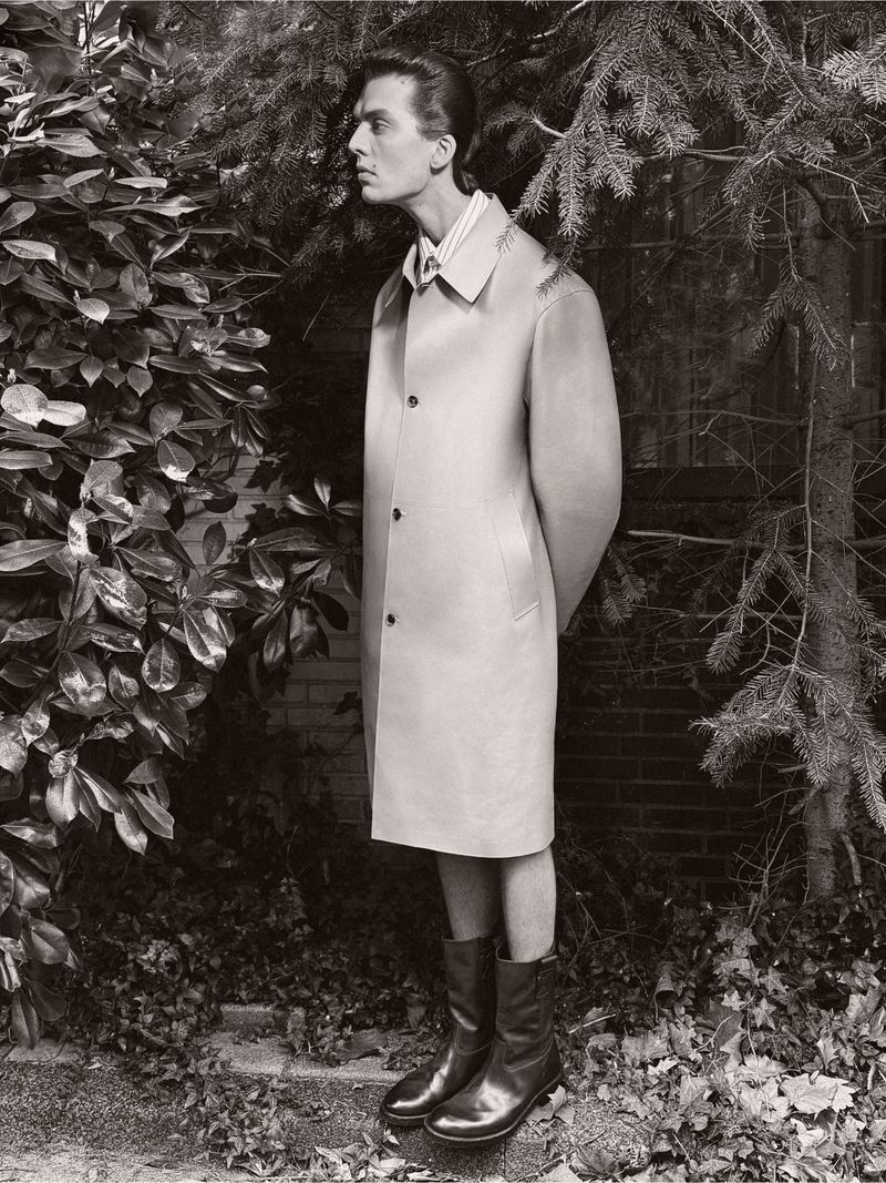Black and white studio shot of a male model posing outdoors in a trench coat and boots, framed by greenery and a brick wall.