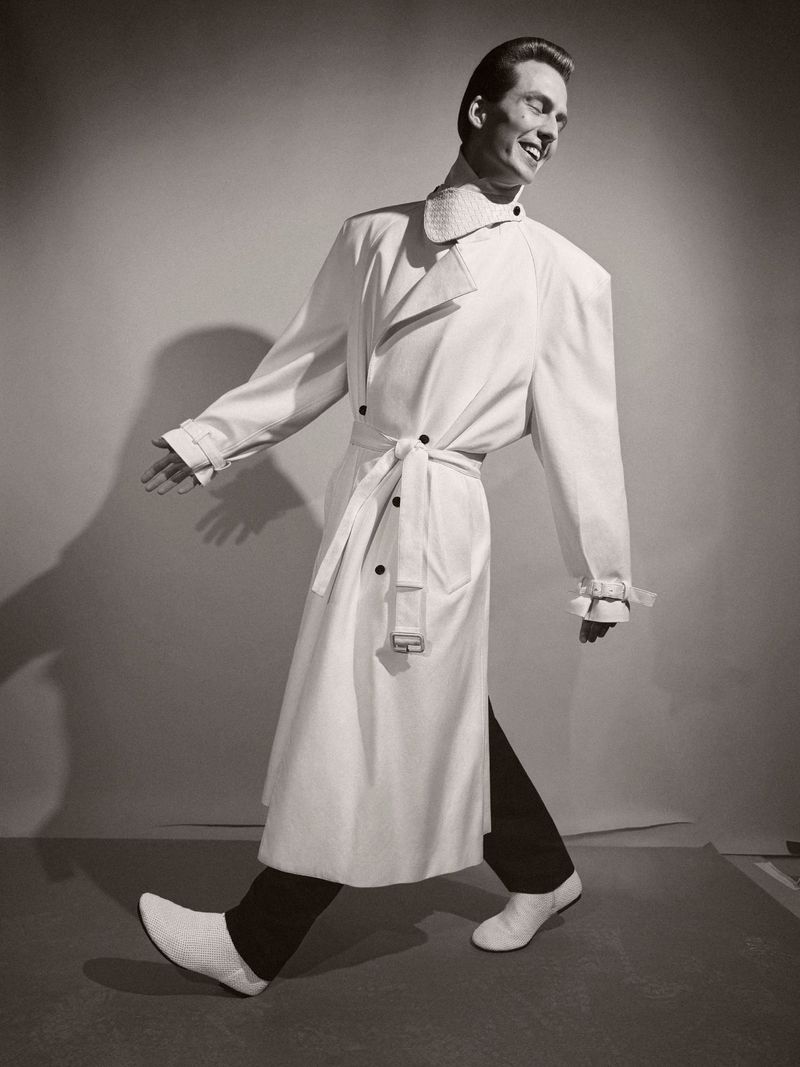 Man in a white trench coat and white shoes strides across a grey background, smiling. Fashion advertisement, studio shot.