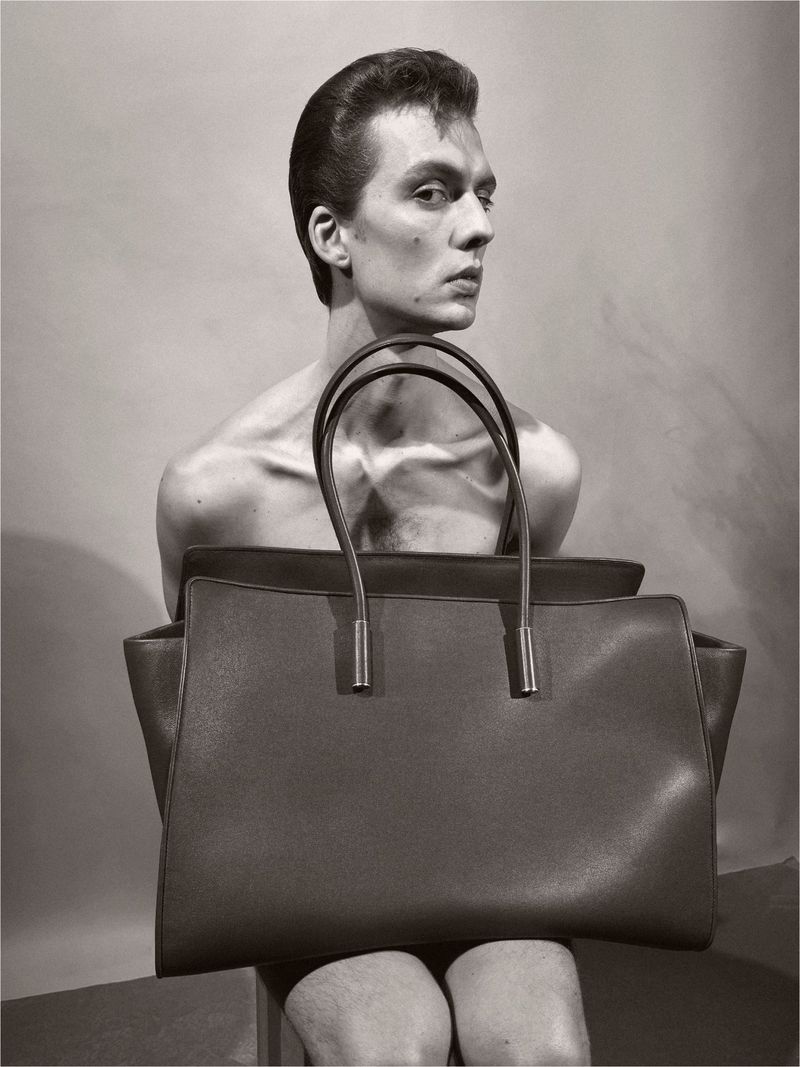 Monochrome studio portrait of a slender, light-skinned adult male with dark hair, posing with a large brown leather tote bag.
