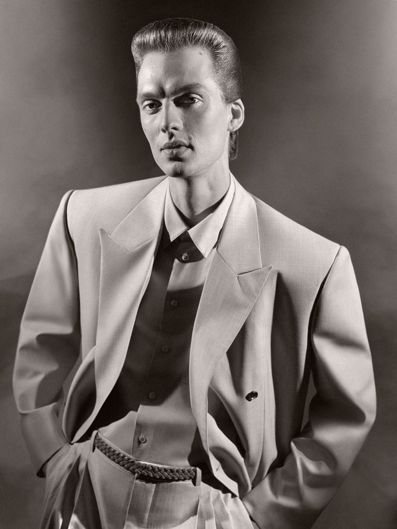 Monochrome portrait of adult male wearing stylish light-colored suit jacket and dress shirt with hands in pockets against dark backdrop.