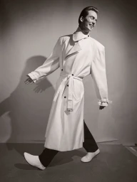 Model Leon Dame smiling while walking in an oversized white belted trench coat against a studio backdrop in black and white.