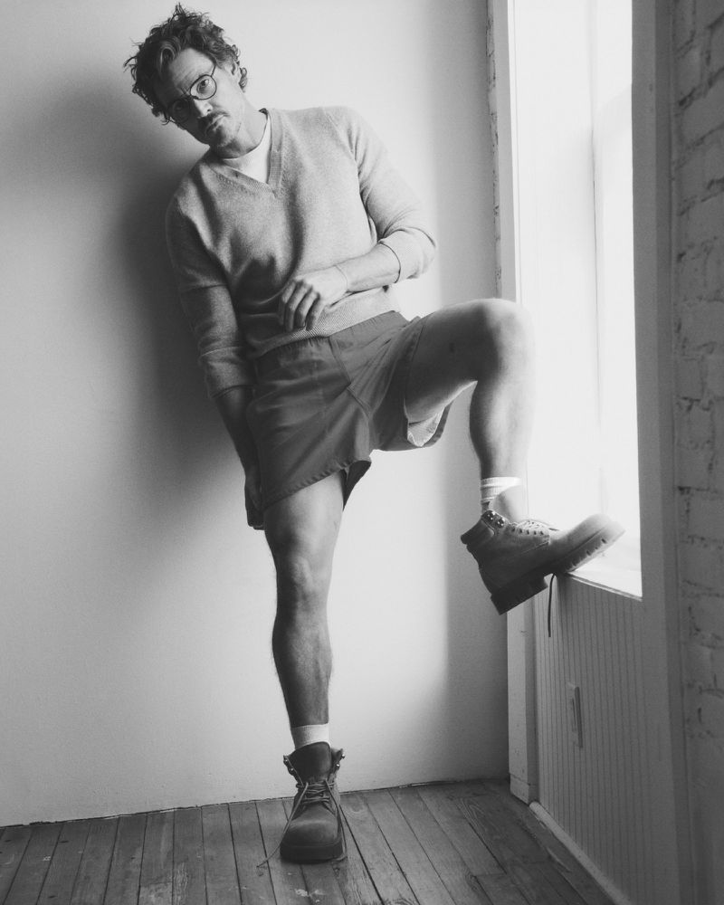 Pedro Pascal wearing a V-neck sweater and shorts, posing with one leg raised against a window sill in a black and white portrait.