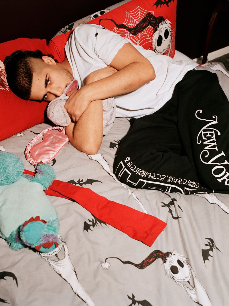 Adult male resting in bed with cartoon-themed bedding, pillows, and sleep mask, creating a casual, indoor scene with a youthful vibe.