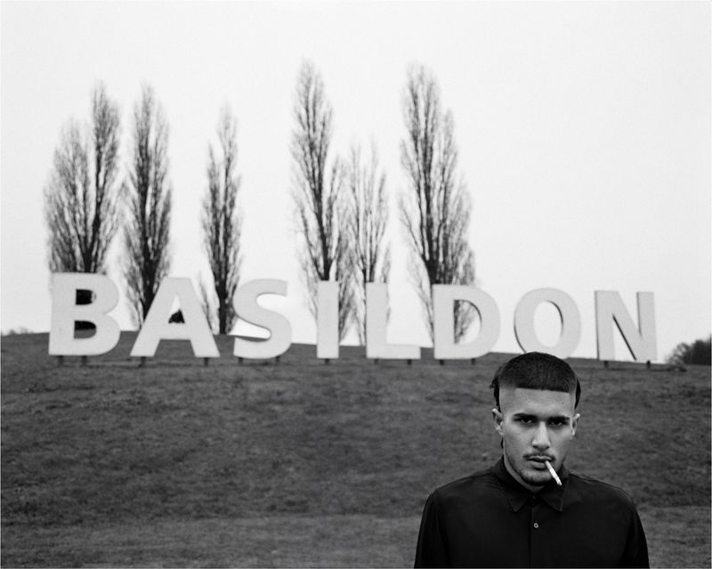 Man with a fade haircut and a cigarette in his mouth stands in front of a sign spelling BASILDON on a grassy hill.