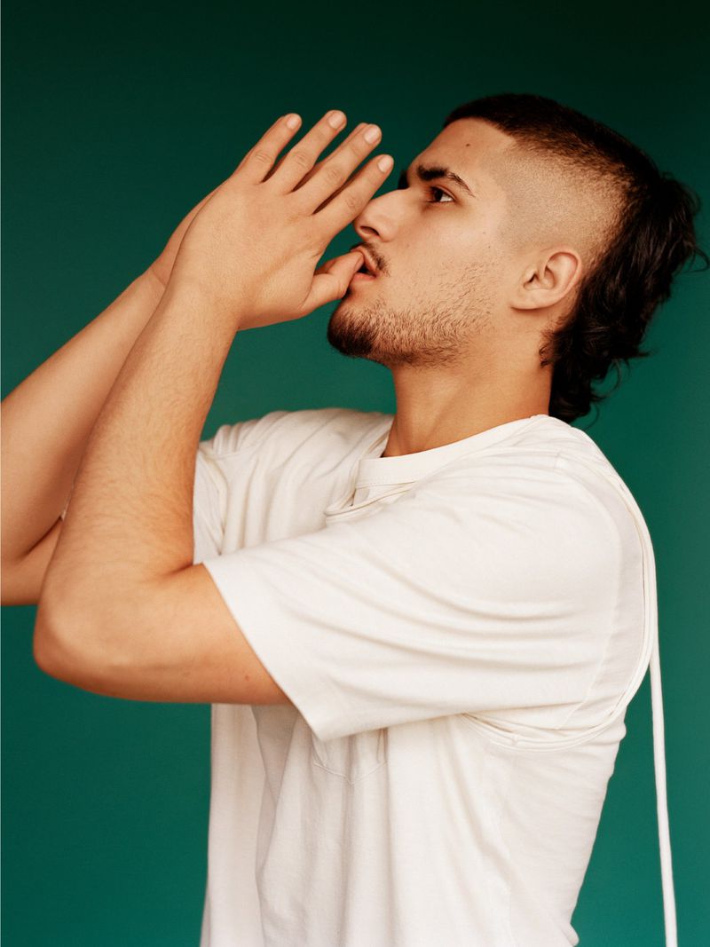 Portrait of a young adult male with a unique mullet hairstyle, wearing a white t-shirt, set against a vibrant teal background.