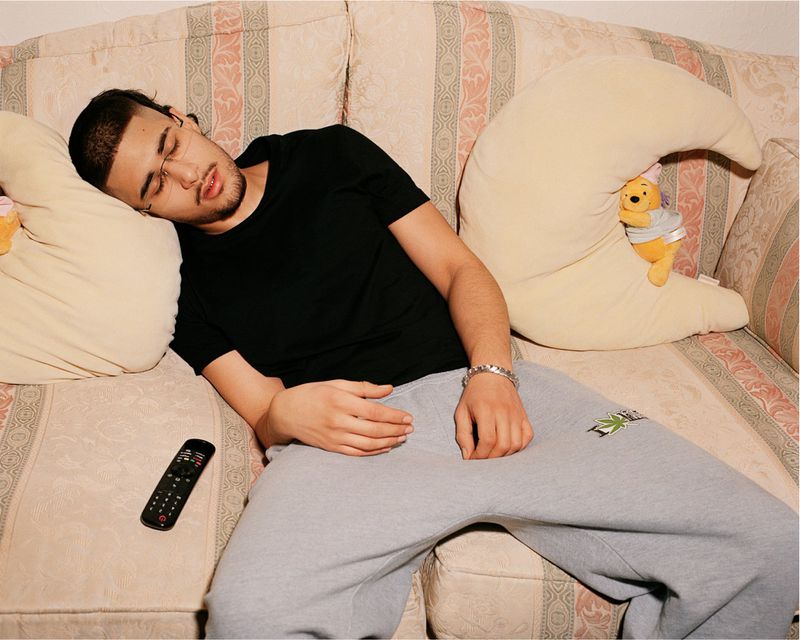 Adult male asleep on a patterned couch with a Winnie-the-Pooh pillow, wearing a black t-shirt and gray sweatpants, with a remote nearby.