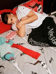 Young man lying in bed holding a small plush toy, wearing a white t-shirt and black sweatpants with New York text