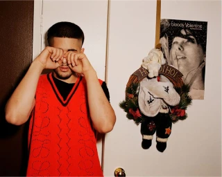 A man wearing a red vest rubs his eyes near a white door with a holiday wreath and a poster.