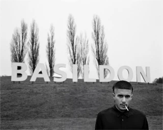 Monochrome portrait of Fakemink smoking a cigarette in front of the large Basildon hillside sign and tall trees.