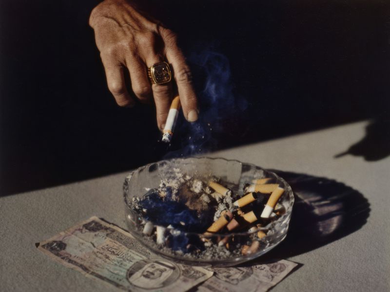 Close-up of hand placing a lit cigarette into an ashtray overflowing with butts and ash, next to currency on a table.
