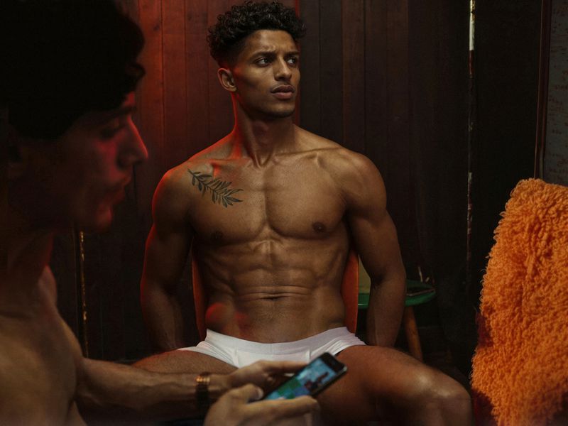 Two shirtless adult men indoors, one seated looking right with a fern tattoo on his shoulder, the other holding a phone, red accent lighting.