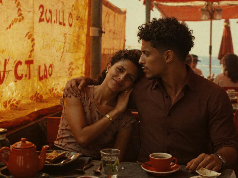 A couple embraces affectionately at a sunlit cafe, sharing tea by a patterned wall, creating a warm and intimate atmosphere.