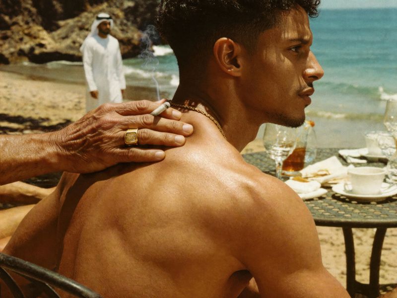 A topless man seated at a table outside, with another man's hand on his back and an Emirati man in the background on a beach.