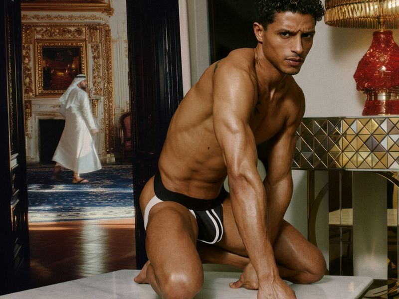 A muscular adult male kneels in a jockstrap in an ornate room with another man walking in the background near a gold mirrored table.