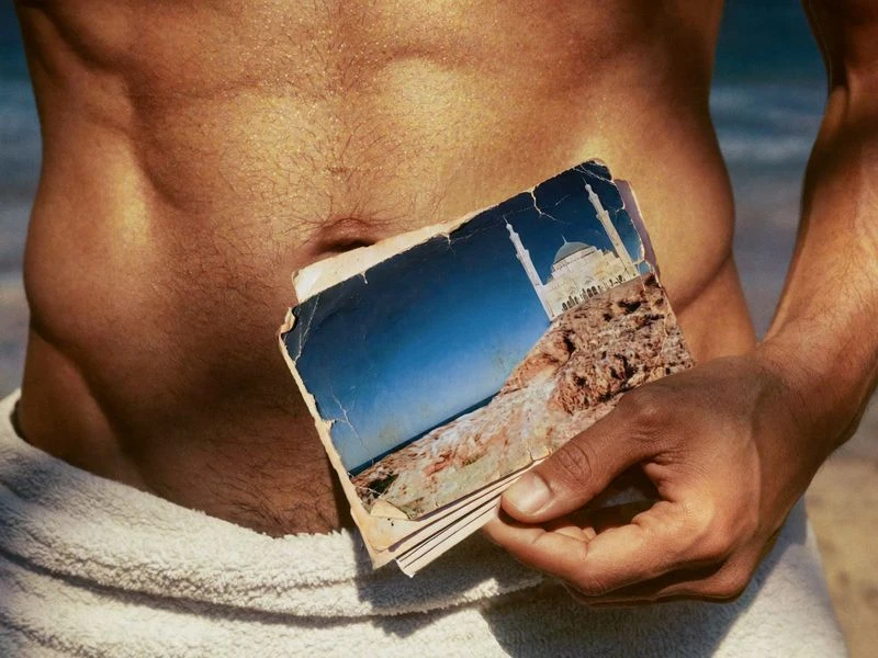Man with a towel around his waist holds up a photograph of a Middle Eastern building on a beach with blue water, sunny day.