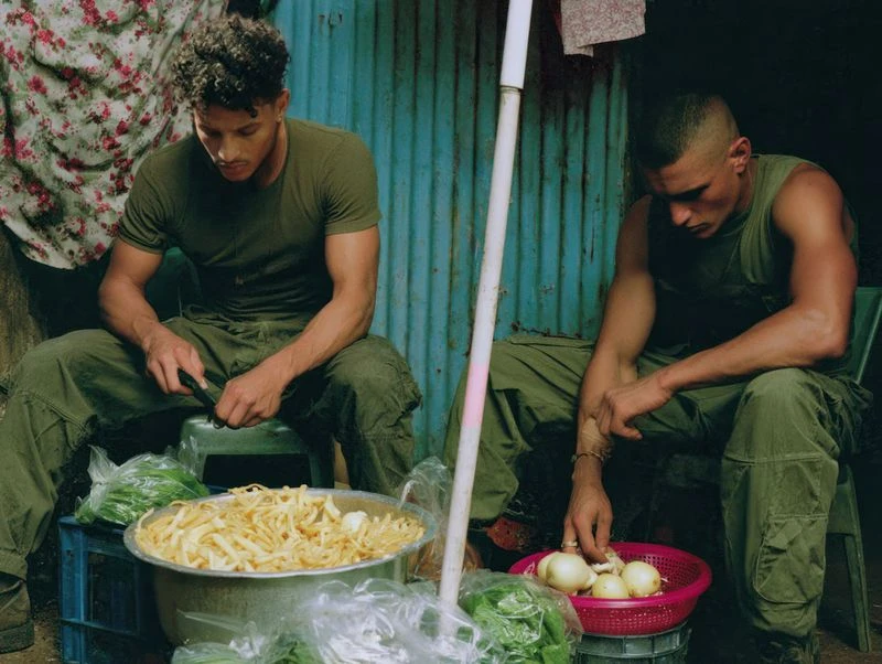 Two men in green work with produce at an outdoor market, using knives to prepare food in bowls, with corrugated metal backdrop.