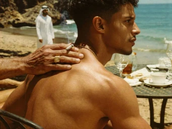 Shirtless young man in profile with a weathered hand resting on his shoulder holding a lit cigarette at a beachside table.