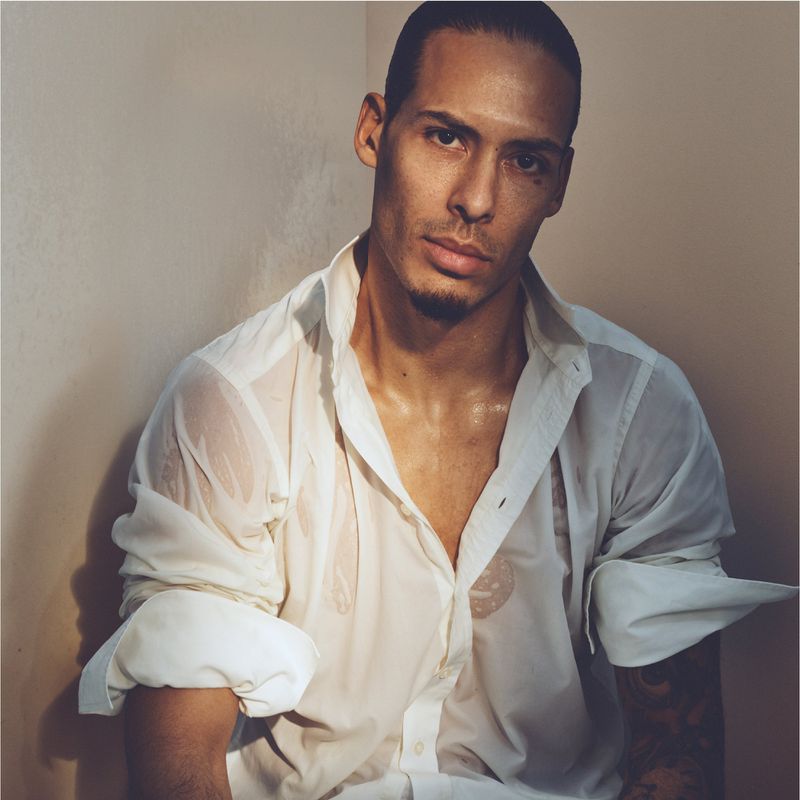 Portrait of a slender adult male with dark hair wearing a wet white collared shirt in a tan-colored indoor setting with natural lighting.