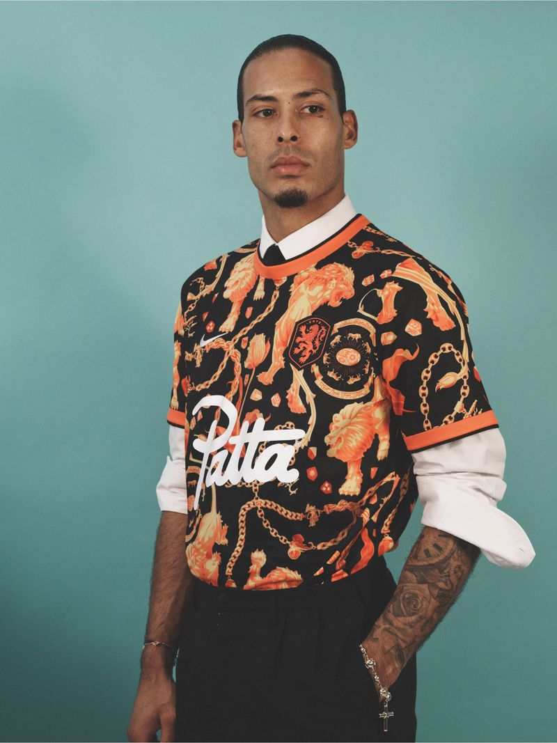 Man in patterned jersey with lions, collars shirt with a tie, wearing bracelets, posing against a teal backdrop in a studio setting.