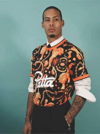 Man in patterned jersey with lions, collars shirt with a tie, wearing bracelets, posing against a teal backdrop in a studio setting.
