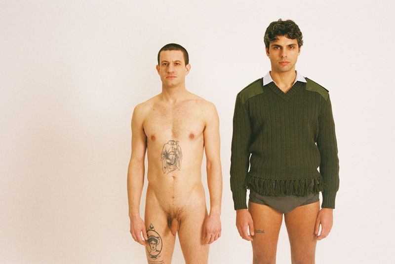 Two men stand side-by-side in a white photo studio. One man is nude with visible tattoos, and the other wears an olive sweater and briefs.