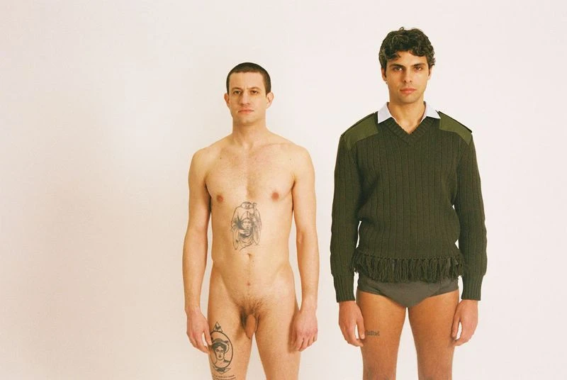 Two men stand side-by-side in a white photo studio. One man is nude with visible tattoos, and the other wears an olive sweater and briefs.
