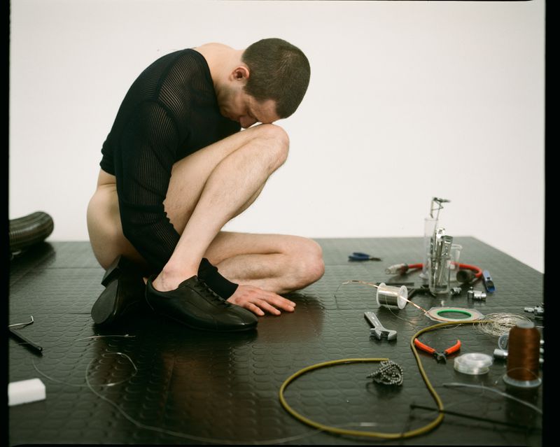 Man in a black shirt is crouching on a black floor surrounded by various tools, wires, and a shoe, set against a plain white background.