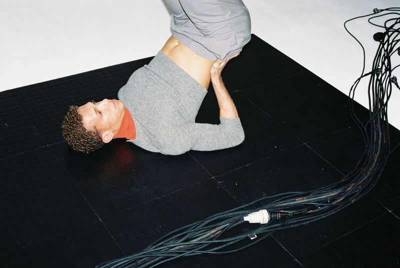 Adult male performs a yoga headstand indoors on a black tiled surface with gray wires and light-gray clothing against a white wall backdrop.