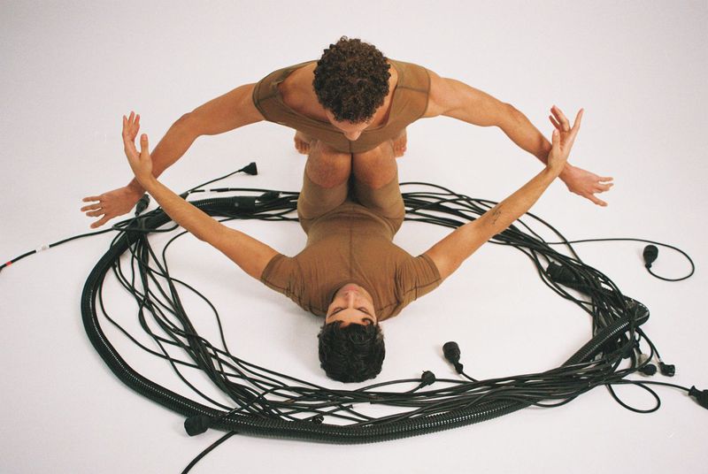 Two adult men wearing brown bodysuits pose with their arms outstretched, surrounded by black electrical cables against a white backdrop.