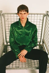 A young man in a shiny green satin zip-up jacket and black trousers sits inside a wire mesh storage crate against a white background.