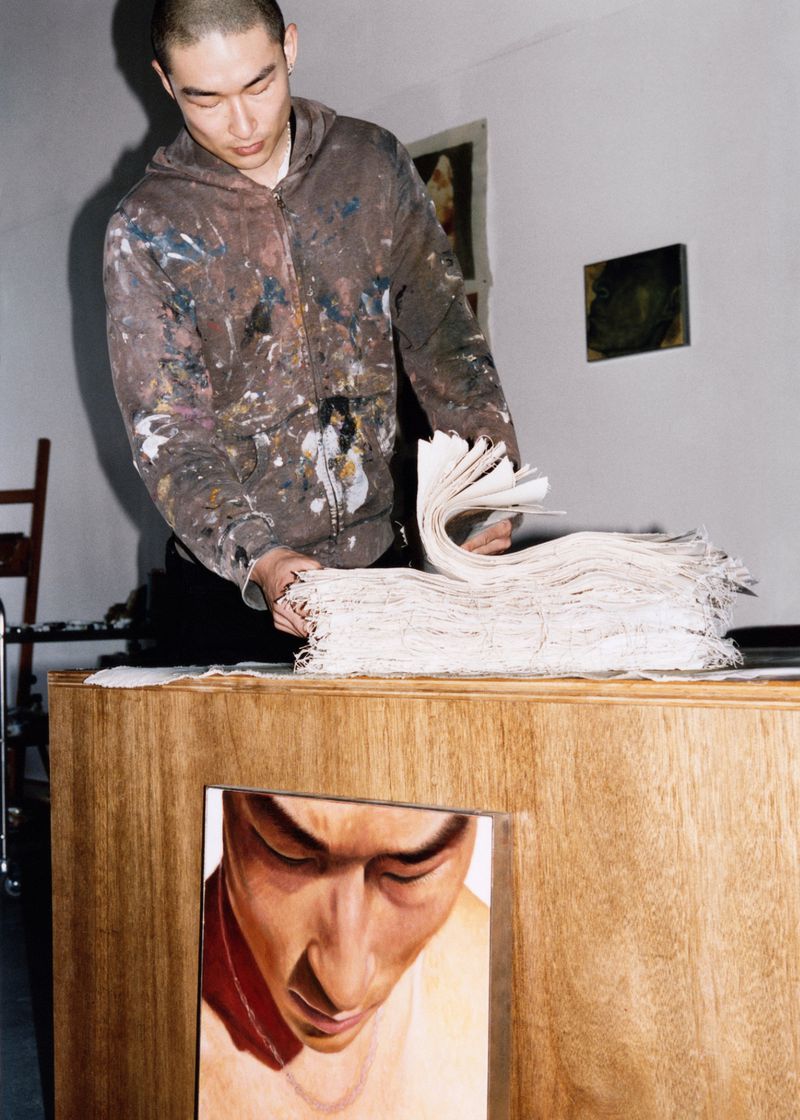 Artist stands at a wooden table, carefully organizing a stack of paper; a painting of a man's face is visible on the front of the table.