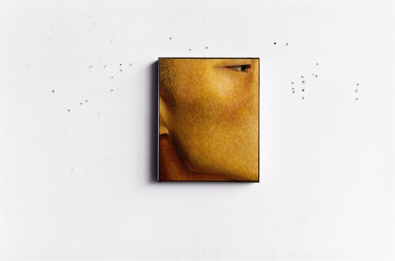 Painting of partial face in a gold frame hanging on a white wall, cropped view of eye, cheek, nose, and forehead. Art installation.