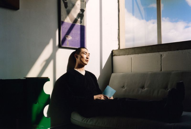 An Asian adult male sits indoors on a light gray sofa in sunlight, holding a blue card, with modern art and a window in the background.