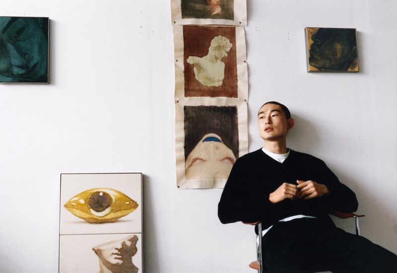 Portrait of an Asian man in a black sweater sitting in a chair in a white room with various paintings on the wall, artful and serene.