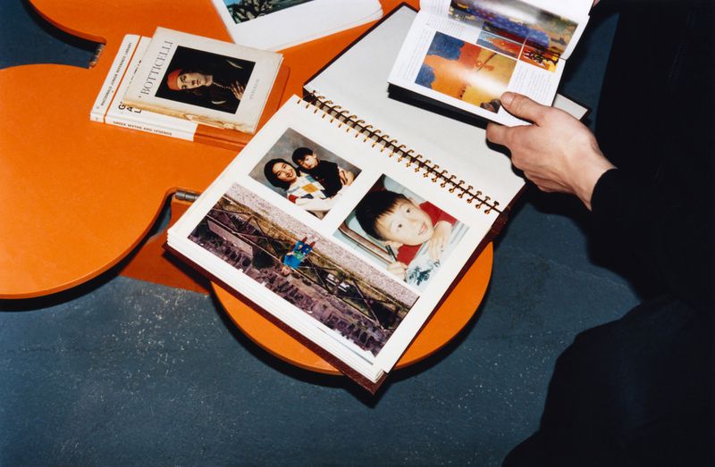 Person holds an open photo album, displaying snapshots of people and scenes on orange table with books and gray concrete floor.