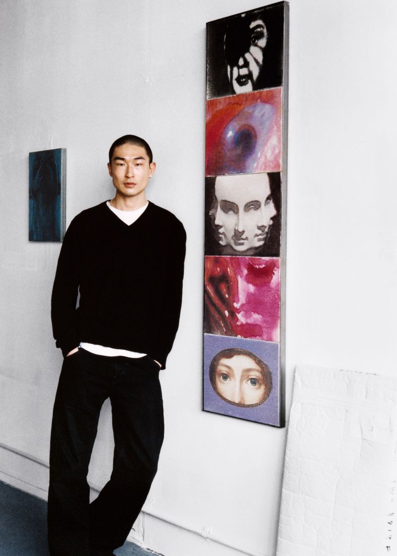 Portrait of a stylish Asian man in black sweater and jeans posing next to tall artwork panels featuring faces in an art gallery.